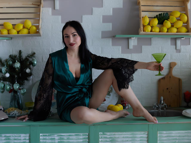 StefanaWinter with black hair, green satin robe with black lace sleeves, sitting on kitchen counter, holding green martini glass, one leg raised, showing her tits, with lemons nearby.