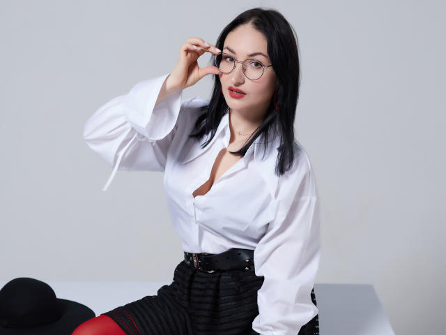 StefanaWinter with glasses, black hair, white shirt, black skirt, red stockings, black belt, black hat on white table, adjusting glasses, sexy, fair skin, red lipstick, cute.