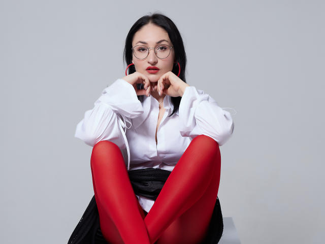 StefanaWinter with red pantyhose, white shirt, black skirt, and glasses, sitting with knees up, hands near face, looking at camera, black hair, pink earbuds.