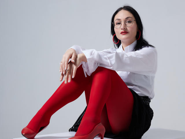 StefanaWinter with red tights, white shirt, black skirt, red heels, glasses, and red earrings, sitting with knees up, hands on knees, looking at camera.