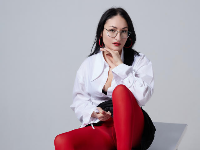 StefanaWinter with black hair, red tights, white shirt, red earrings, glasses, sitting, hand near face, black skirt, small tits, red lips, looking at camera.