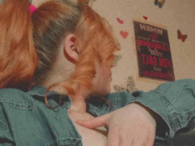 SpicyGrace, con i capelli rossi raccolti in due codini, indossa una camicia di jeans verde, con una mano vicino al seno, che guarda di lato, in una stanza con decorazioni a forma di cuore e un poster..