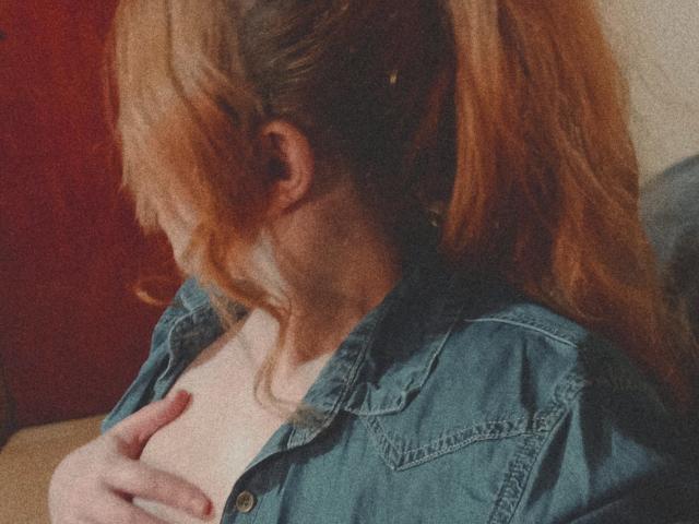 SpicyGrace, con i capelli rossi raccolti in una coda di cavallo, indossa una camicia di jeans blu e tocca i suoi seni nudi con una mano, guardando di lato. Le sue dita afferrano delicatamente il suo seno..