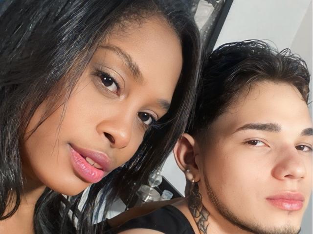 Damaris has glossy pink lips and dark straight hair, next to Cris with short dark hair, a goatee, and a tattoo on his neck. Both have brown eyes and black shirts.
