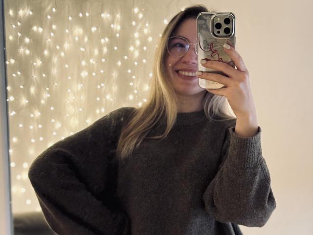 MiaFlirt, avec des lunettes, des cheveux blonds, souriante, portant un pull gris, tenant un téléphone avec une coque rouge à motifs, prenant un selfie devant un miroir. Les seins ne sont pas visibles..