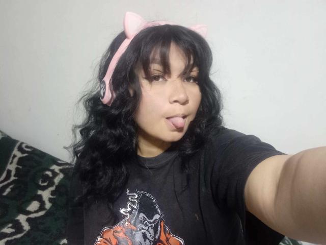 LillyMayer, con cabello negro ondulado, orejas de gato rosas y una camiseta negra, saca la lengua y se toma un selfie con su brazo derecho. Sus glúteos no son visibles..