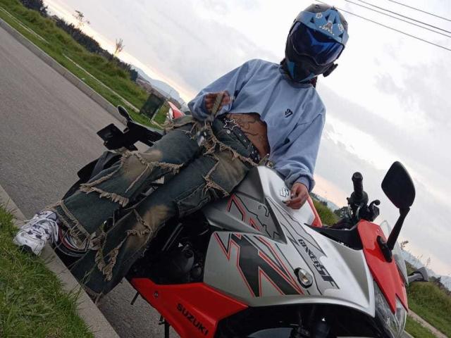 SharlotteX, wearing a blue helmet and gray hoodie, sits on a red Suzuki motorcycle with ripped jeans and white sneakers, looking down with a slight smile.
