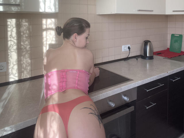 LinaShy with back to camera, pink strapless top, red panties, tattoo on right butt cheek, leaning on kitchen counter, black stove, stainless kettle, beige tiles.