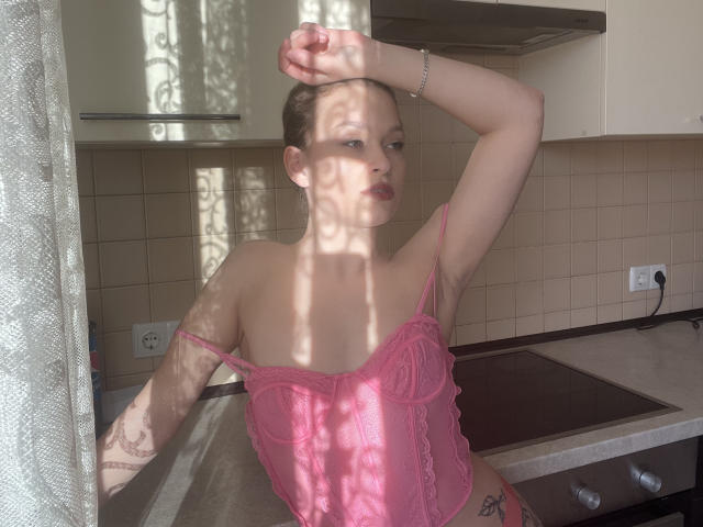 LinaShy with bare tits in pink lace lingerie, arm raised, sunlight casting shadows, standing in kitchen, light skin, brown hair, tattoo on arm, looking away.