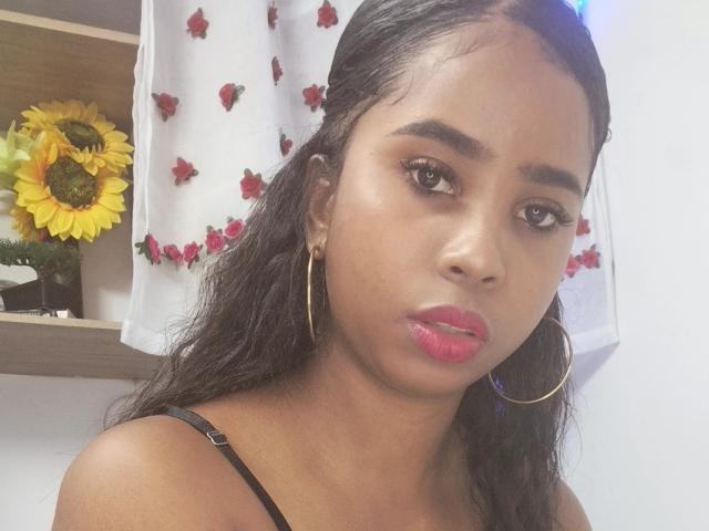 LiaLuz with medium brown skin, long black hair, hoop earrings, pink lipstick, and brown eyes, wearing a black spaghetti strap top, looking serious at the camera.