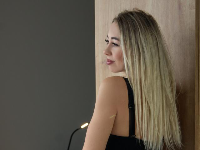 MargaritaBeauty with long blonde hair, black dress, smiling sideways, leaning against wooden wall, small bandage on upper arm, lamp in background, soft lighting.