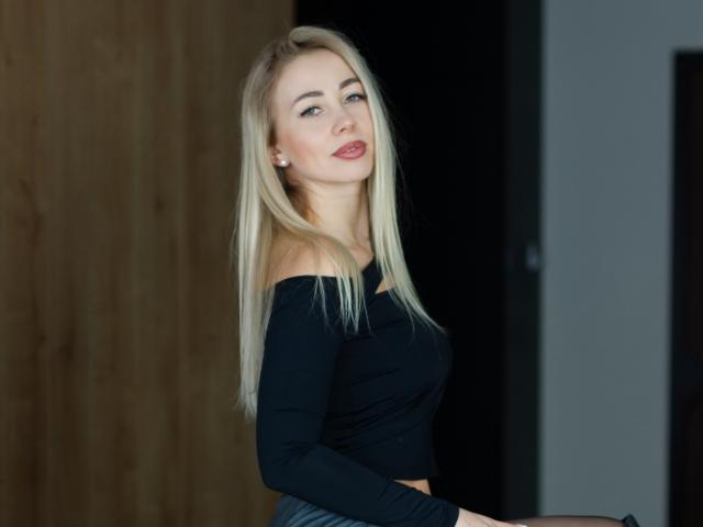 MargaritaBeauty with blonde hair, black off-shoulder top, and subtle makeup, showing her tits and ass, looking seductive with a slight smile.