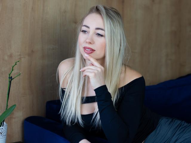 MargaritaBeauty with blonde hair, black off-shoulder top, and pink lips, smiling slightly, hand near mouth, sitting on blue couch, wooden wall background, small green plant left.