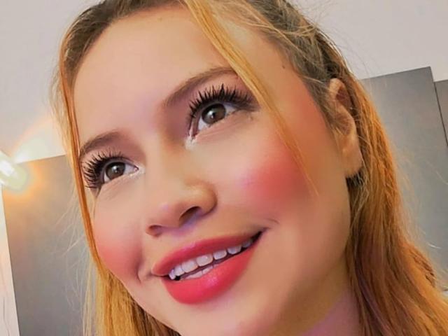 KeniaBlondie's face with light brown hair, pink blush, and red lipstick, smiling with bright eyes and long lashes, has a cute, pretty, sexy look.