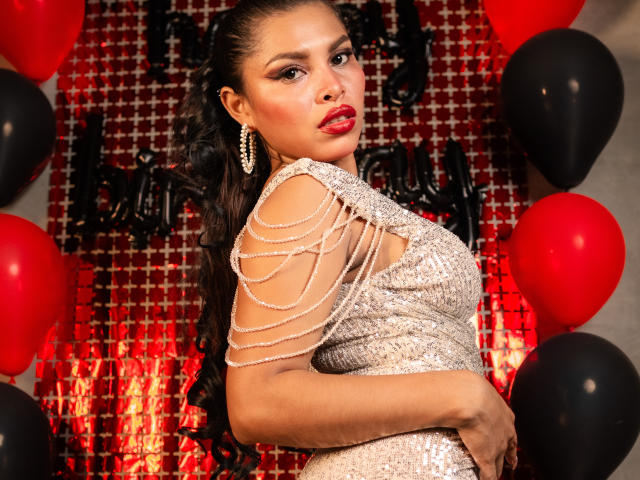 MarceBetancur in a sparkly, sleeveless dress with chain-like straps, red lipstick, and long, curly black hair, surrounded by red and black balloons, looking over her shoulder.