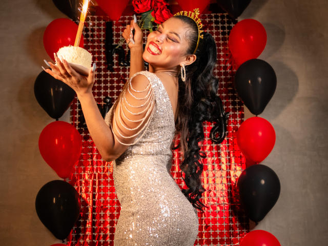 MarceBetancur in a sparkly silver dress, holding a cake with a candle, smiling with red lipstick, black hair in a ponytail, gold headband, surrounded by red and black balloons.