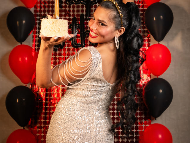 MarceBetancur with long black hair, glittery silver dress, and diamond earrings, holding a fluffy white cake, smiling, surrounded by red and black balloons.