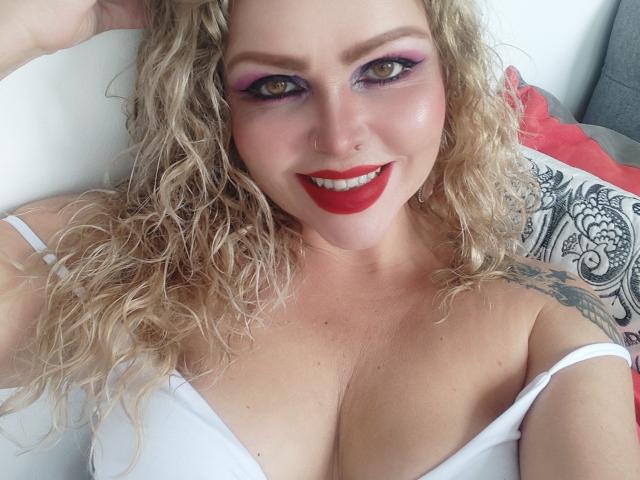 AnnaRichie with curly blonde hair, red lipstick, purple eyeshadow, smiling, wearing a white strap dress, showing her large boobs, and a tattoo on her right shoulder.