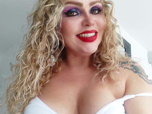 AnnaRichie with curly blonde hair, purple eyeshadow, red lipstick, white strapless top, showing her big tits, nose ring, and tattoo on her right shoulder, smiling.