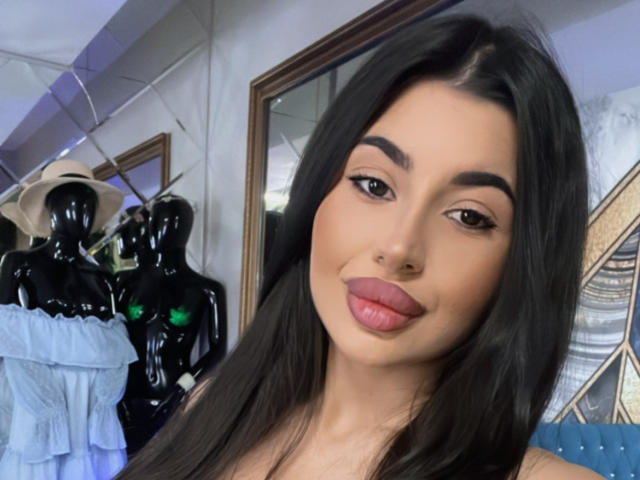 LatinaLoco with long black hair, full pink lips, and dark eyebrows, smiling in a selfie. She's in front of glossy black mannequins wearing blue dresses and hats.