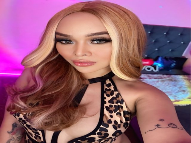 UrDreamAsianTransPinay69 with blonde hair, heavy makeup, and leopard print halter top, showing off her tits and tattoos, sitting in a pink-lit room.