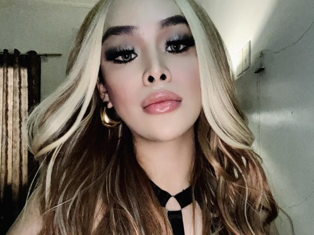 UrDreamAsianTransPinay69 with long blonde and brown wavy hair, thick black eyeliner, glossy pink lips, and large gold hoop earrings, wearing a black choker.