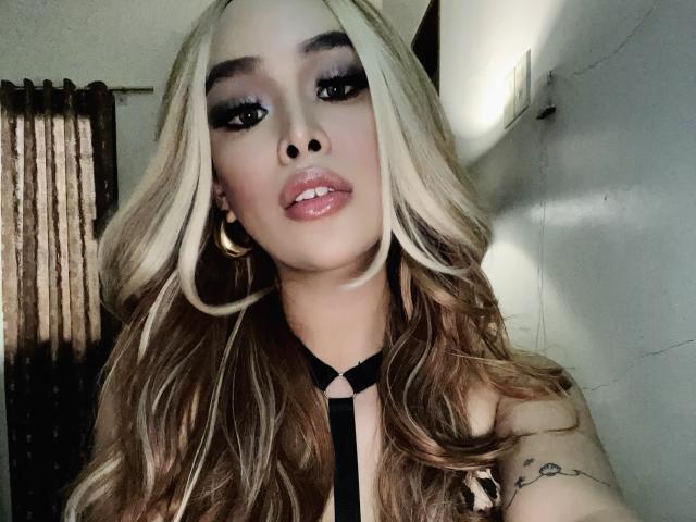 UrDreamAsianTransPinay69 with blonde and brown wavy hair, dark eyeliner, glossy pink lips, gold hoop earrings, black strap top, tattoo on right arm, looking at camera, in dim light.
