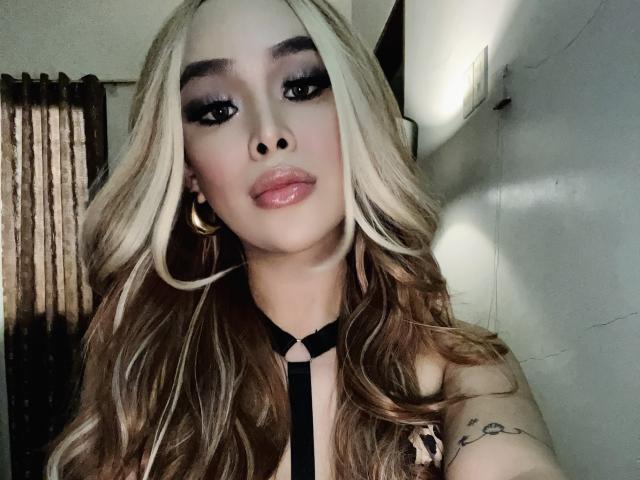 UrDreamAsianTransPinay69 with long blonde hair, glossy lips, black eyeliner, gold hoop earrings, black strappy top, tattoo on right arm, looking at camera with soft lighting.