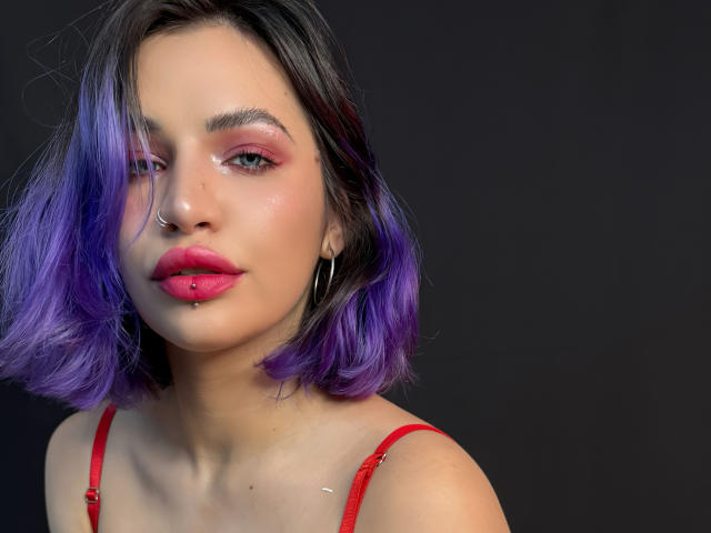 BriannaAngel69 has purple-dyed hair, pink eyeshadow, red lipstick, and a nose ring. Her face is partially hidden by a gray bar. She has a nose piercing and hoop earrings.