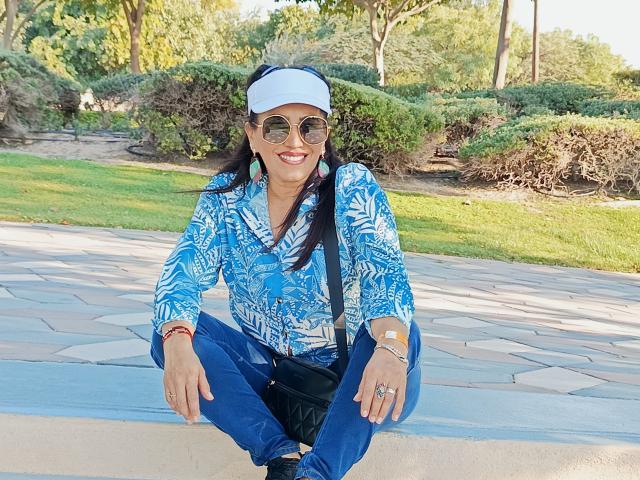 MelissaDelici, avec de longs cheveux noirs, portant une casquette blanche, une chemise à motifs bleus, un pantalon bleu et des lunettes de soleil, assise sur une surface en pierre, souriante, avec un sac noir sur son épaule..