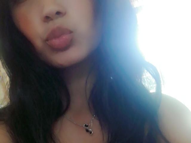 EmaRelling's pouty, glossy lips are slightly up, showing off her full, dark brown, wavy hair and a simple necklace with small black and silver charms.