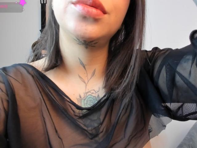 KayleeM with light skin, black sheer top, visible tattoo of a flower on her chest, brown hair, and slightly parted lips. Her tits are partially covered by the sheer fabric.