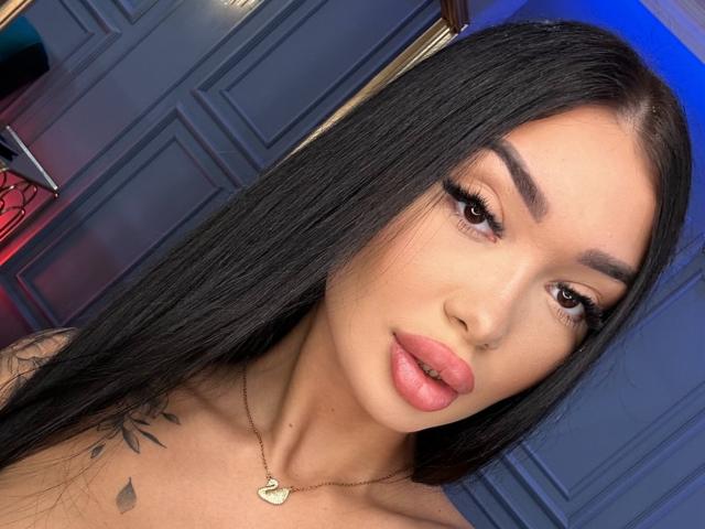 SashaFerrera with long black hair, full pink lips, brown eyes, and a gold necklace. She has a tattoo on her right shoulder and is looking at the camera.