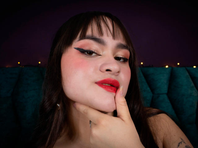 LailaDurmont has dark brown hair, red lipstick, and black winged eyeliner. She's touching her chin with a finger, showing a tattoo on her right hand.