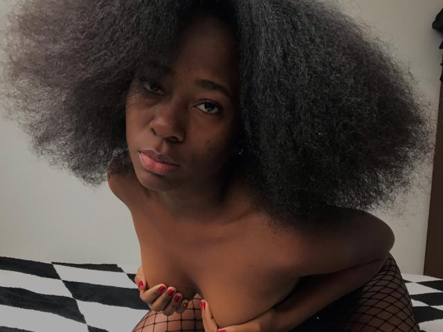 VioletGraice with dark curly hair, topless, wearing fishnet stockings, covering small tits with hands, red nails, pouty expression, looking up, in a black-and-white striped bed.
