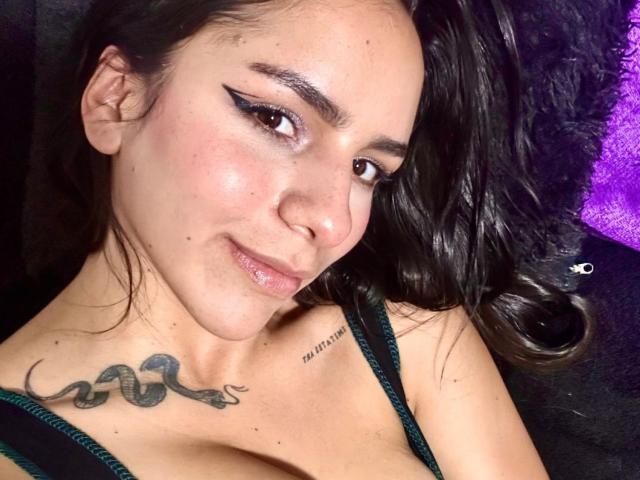LizzyGrant69 with dark hair, black eyeliner, and a snake tattoo on her chest, smiling at the camera, wearing a green strap dress, showing her tits.