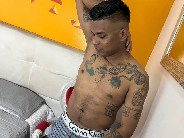 SamSexy69, sin camisa, tatuado, vistiendo ropa interior Calvin Klein, de pie en un dormitorio, con un brazo levantado, cabello negro corto, mirando hacia abajo, con tatuajes en el pecho y el brazo, una cama gris con un cojín rojo, una pared amarilla..