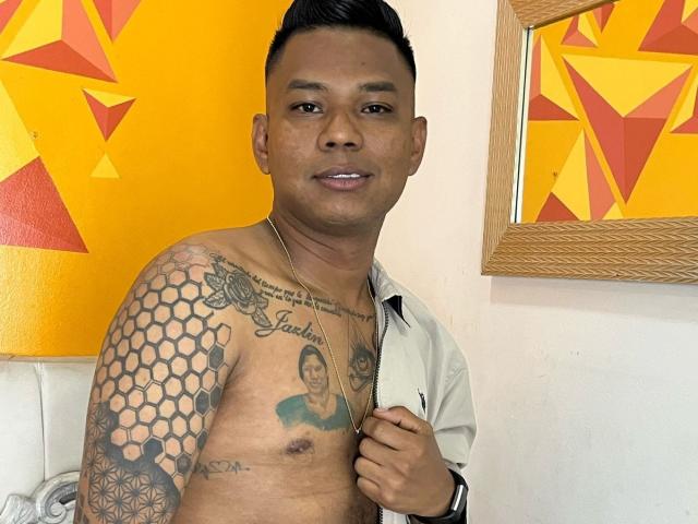 SamSexy69 senza camicia, con tatuaggi, che tiene una camicia, capelli neri corti, pelle marrone, petto piccolo, tatuaggio esagonale sulla spalla destra, tatuaggio a forma di ritratto sul petto, sorridente, parete giallo-arancione, cornice di legno..