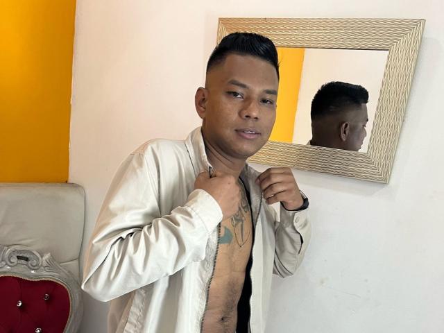 SamSexy69, senza camicia con una giacca bianca, tiene il colletto, capelli neri corti, tatuaggio sul petto, riflesso nello specchio, parete gialla, letto rosso e bianco, espressione seria, pelle scura..