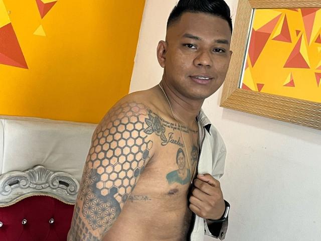 SamSexy69, senza camicia, con tatuaggi esagonali e geometrici sulla spalla destra, capelli neri corti, pelle color nocciola, sorridente, che tiene una giacca parzialmente aperta, opere d'arte a parete rosse e gialle..