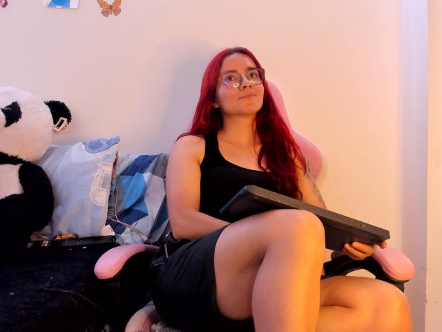 EimmyHoskX with red hair, glasses, black tank top, and black skirt, sitting on pink-and-black chair, holding game controller, legs crossed, large ass, small tits, looking up.