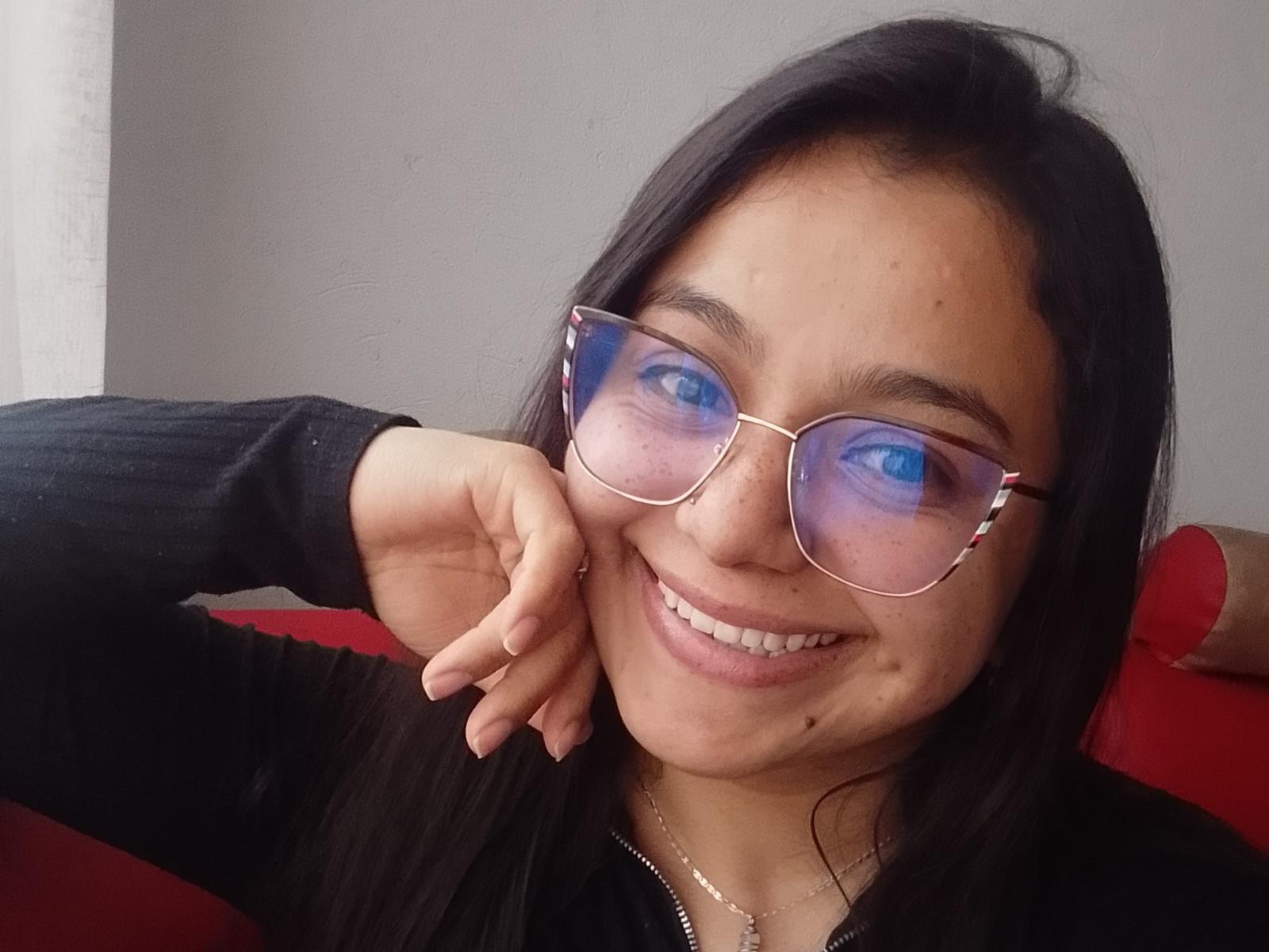 EvaHobson with blue reflective sunglasses, black hair, smiling, wearing a black top, hand resting on cheek, showing her white teeth and a small beauty mark on her right cheek.