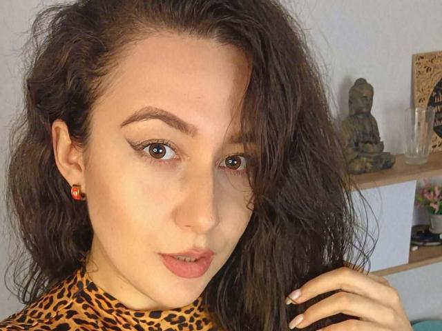 MabyLegrand with wavy brown hair, wearing a leopard print top, has brown eyes, red earrings, and slightly parted lips, looking at the camera with a subtle smile.