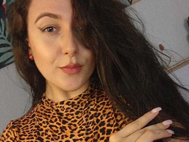MabyLegrand with long brown hair, wearing a leopard print top, has full lips and red earrings, showing a slight smile, with fingers near her face, brown eyes.