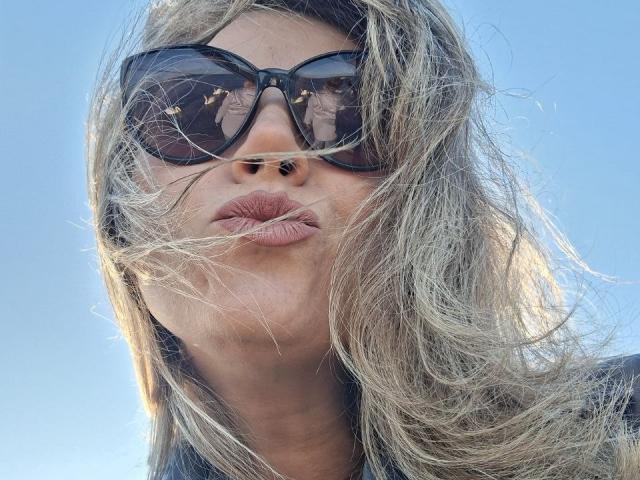 CandyLadyZ with messy blonde hair, pouty pink lips, and big black sunglasses. Her nose is pierced, and she's looking down. Her face is slightly blurred by wind.