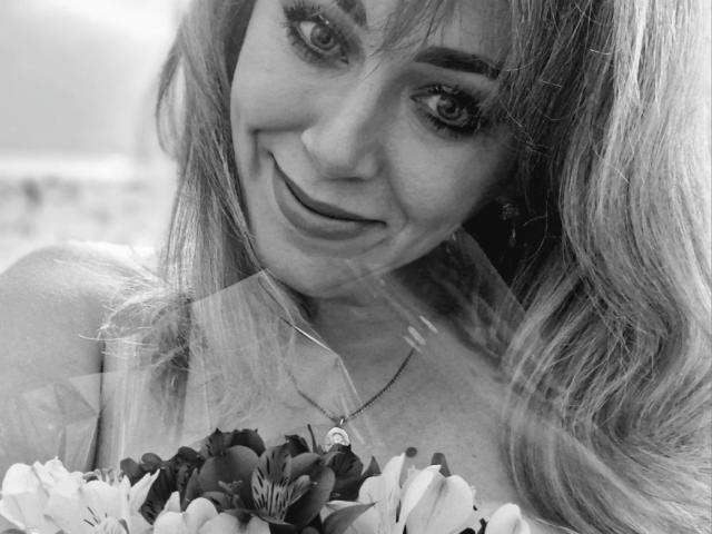 Black-and-white photo of CandyLadyZ with long hair, smiling at the camera, holding a bouquet of flowers, wearing a necklace with a heart pendant, showing her tits.