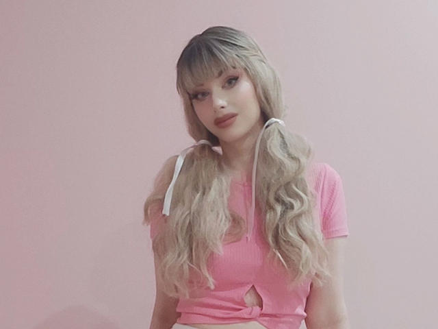 ChloeKiss-hot with long, wavy blonde hair in pigtails, wearing a pink ribbed top with a keyhole cutout, pink background, looking at camera with a slight smile.