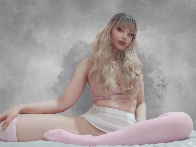 ChloeKiss-hot with long blonde hair, pink lace bra, white skirt, and pink thigh-highs, sitting on a bed, showing her tits and pussy, looking at the camera.