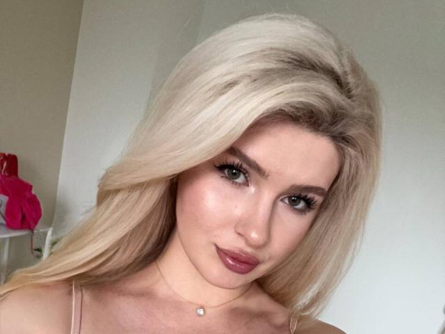 CocoSecrets69 has blonde hair, green eyes, and pink lipstick. She's wearing a sheer top with a small pendant necklace. Her tits are slightly visible. She's giving a sexy look.