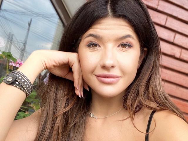 Amylin69 with brown eyes, long brown hair, and light skin, smiling, wearing a black spaghetti strap top, silver necklace, and multiple bracelets, resting her head on her hand.